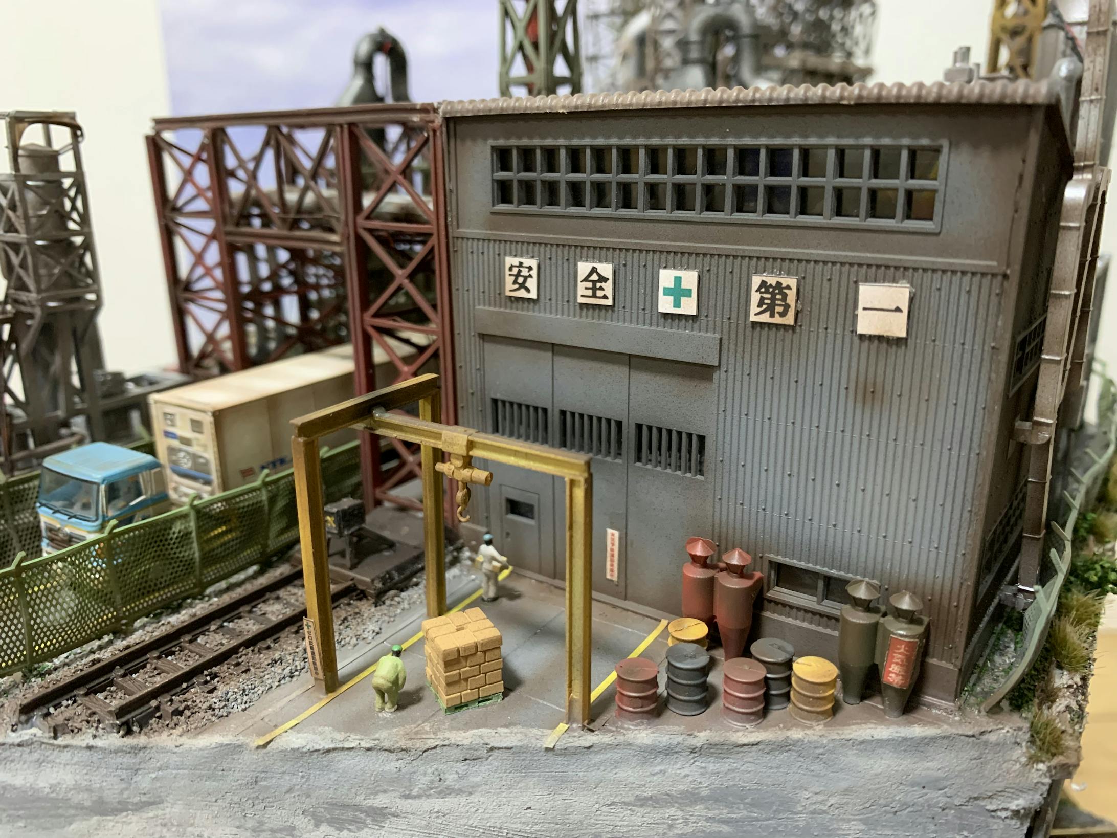 工事周りの表現をどうしていくか考えました。とにかく専用線とかは好きなのでムダに作ってみました。専用線の奥は、車両、歩行者が通るため、安全上フェンスをつけました。クレーンは専用線で運ばれて来た荷物を運搬するために必要なので設置しました。また、ダンボールに入った荷物を作業員が工事に移動させています。ドラム缶の燃料は、工事の稼働に必要です。そんなイメージのもと、配置していきました。